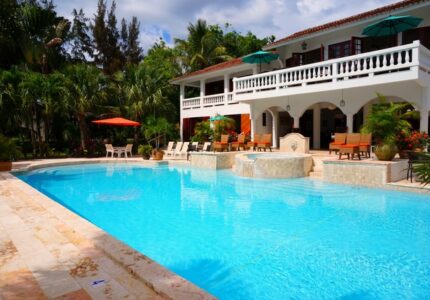 A luxurious white villa with an arched balcony overlooks a large, inviting swimming pool. The pool is surrounded by sun loungers and umbrellas, amidst lush tropical greenery, creating a serene and relaxing atmosphere.