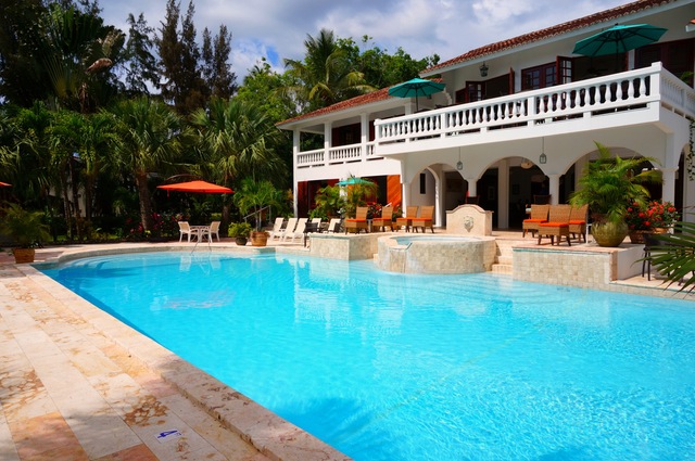 A luxurious white villa with an arched balcony overlooks a large, inviting swimming pool. The pool is surrounded by sun loungers and umbrellas, amidst lush tropical greenery, creating a serene and relaxing atmosphere.