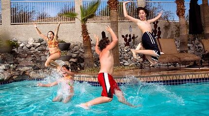 Four children joyfully jump into a swimming pool. One is mid-air with arms raised, another splashes water, and two others leap with excitement. Lounge chairs and tropical plants can be seen poolside on a sunny day.