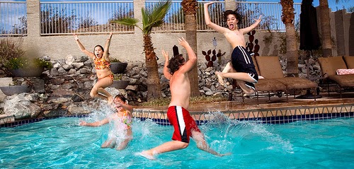 Four children joyfully jump into a swimming pool. One is mid-air with arms raised, another splashes water, and two others leap with excitement. Lounge chairs and tropical plants can be seen poolside on a sunny day.