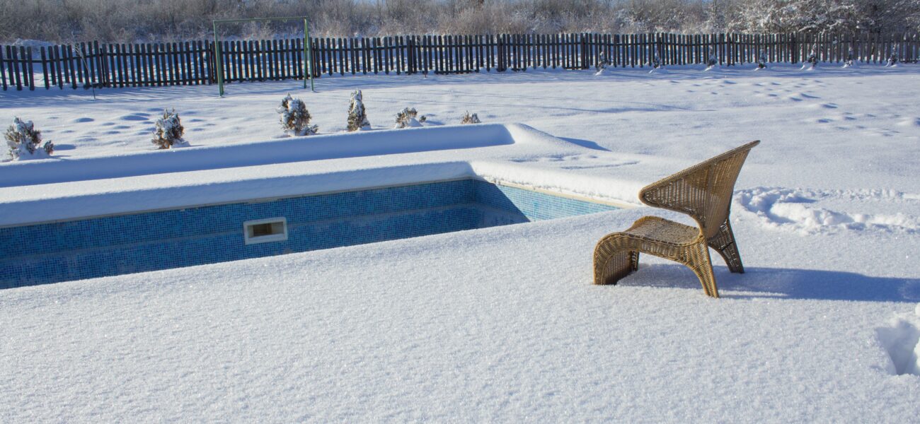 A wicker chair is partially buried in snow beside an empty, snow-covered swimming pool. The surrounding area is blanketed in snow, with a wooden fence and leafless trees in the background under a clear blue sky.