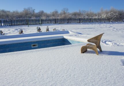 A wicker chair is partially buried in snow beside an empty, snow-covered swimming pool. The surrounding area is blanketed in snow, with a wooden fence and leafless trees in the background under a clear blue sky.