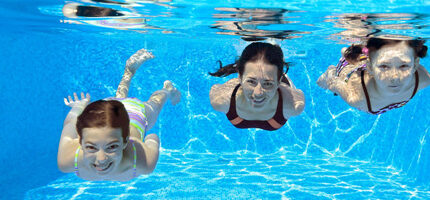 Three children swimming underwater in a pool, smiling at the camera. They are wearing colorful swimwear, and sunlight creates patterns on the pool walls and floor.