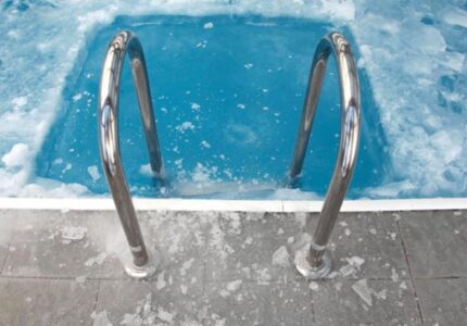 Metal pool steps descend into a swimming pool partially covered with ice. The surrounding tiles are also frosted, indicating cold weather. The water appears icy blue, and shards of ice float on the surface.