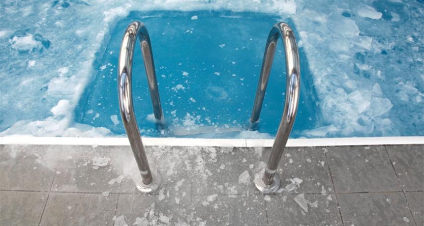 Metal pool steps descend into a swimming pool partially covered with ice. The surrounding tiles are also frosted, indicating cold weather. The water appears icy blue, and shards of ice float on the surface.