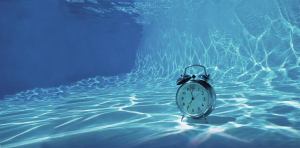 A retro alarm clock sits underwater, illuminated by sunlight creating wavy patterns on the pool floor. The clock, with its metal bells, is fully submerged, adding a surreal, dreamlike quality to the scene.