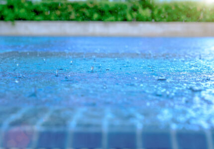 Close-up of raindrops splashing on a blue-tiled surface, with a green hedge and a concrete wall blurred in the background. The water creates a shimmering effect in the sunlight.