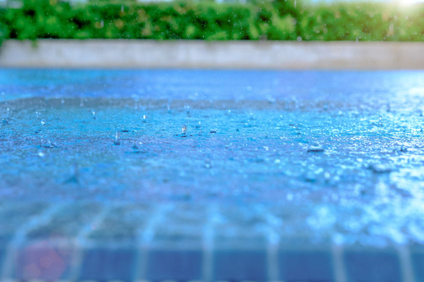 Close-up of raindrops splashing on a blue-tiled surface, with a green hedge and a concrete wall blurred in the background. The water creates a shimmering effect in the sunlight.