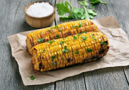 Two grilled corn cobs garnished with herbs sit on brown paper on a wooden table. A small wooden bowl of coarse salt and some parsley leaves are in the background.