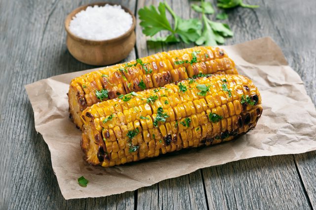 Two grilled corn cobs garnished with herbs sit on brown paper on a wooden table. A small wooden bowl of coarse salt and some parsley leaves are in the background.