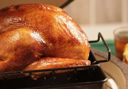 A golden-brown roasted turkey rests on a rack inside a baking pan. In the background, there are glasses of iced tea and a bowl filled with vegetables and lemon slices. The scene suggests a festive or holiday meal setting.