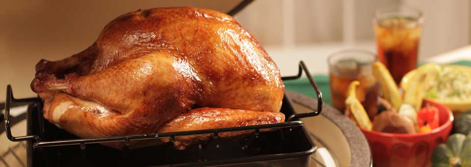 A golden-brown roasted turkey rests on a rack inside a baking pan. In the background, there are glasses of iced tea and a bowl filled with vegetables and lemon slices. The scene suggests a festive or holiday meal setting.