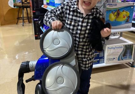 A young child with curly hair and glasses smiles while holding parts of a pool vacuum in a store. Hes wearing a black and white checkered shirt and jeans. The store has a variety of pool supplies and products on shelves behind him.