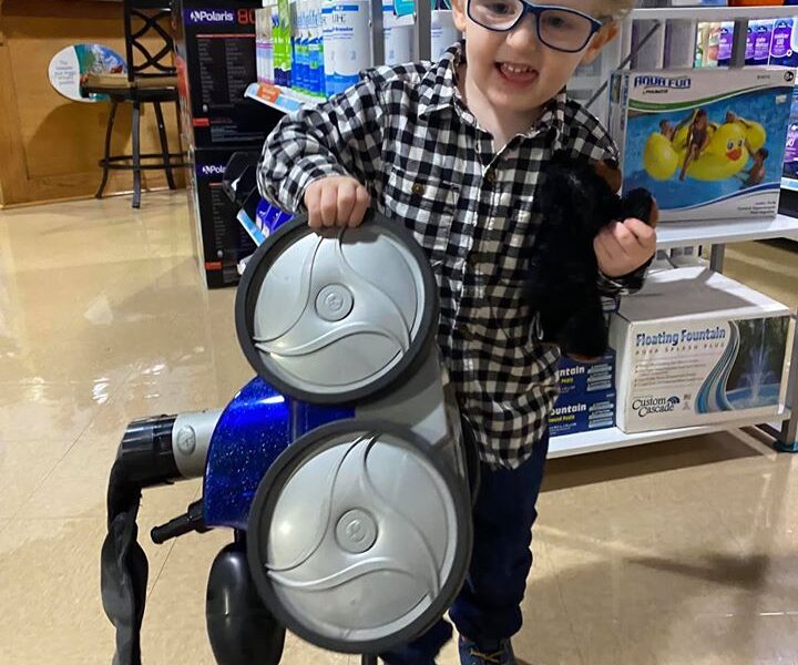 A young child with curly hair and glasses smiles while holding parts of a pool vacuum in a store. Hes wearing a black and white checkered shirt and jeans. The store has a variety of pool supplies and products on shelves behind him.