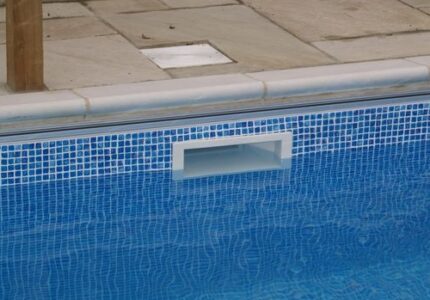 A close-up of a swimming pool edge with small blue tiles and a rectangular white pool skimmer. The pool water is calm, reflecting the tiles. Stone paving surrounds the pool area.