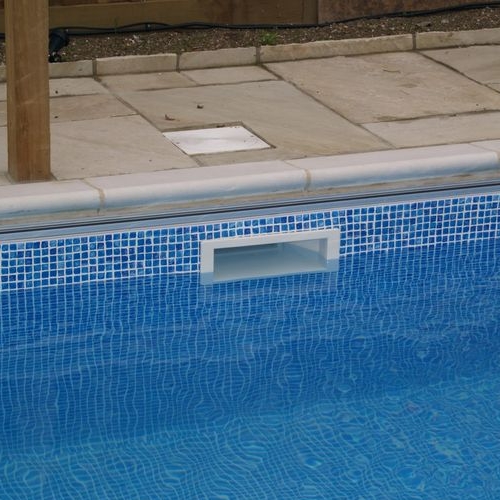 A close-up of a swimming pool edge with small blue tiles and a rectangular white pool skimmer. The pool water is calm, reflecting the tiles. Stone paving surrounds the pool area.