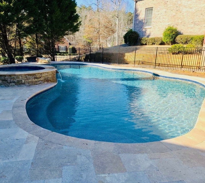 A backyard swimming pool with clear blue water, surrounded by a stone deck. The pool is bordered by a wrought-iron fence, and there are trees and a brick house in the background. Sunlight casts shadows on the pool surface.