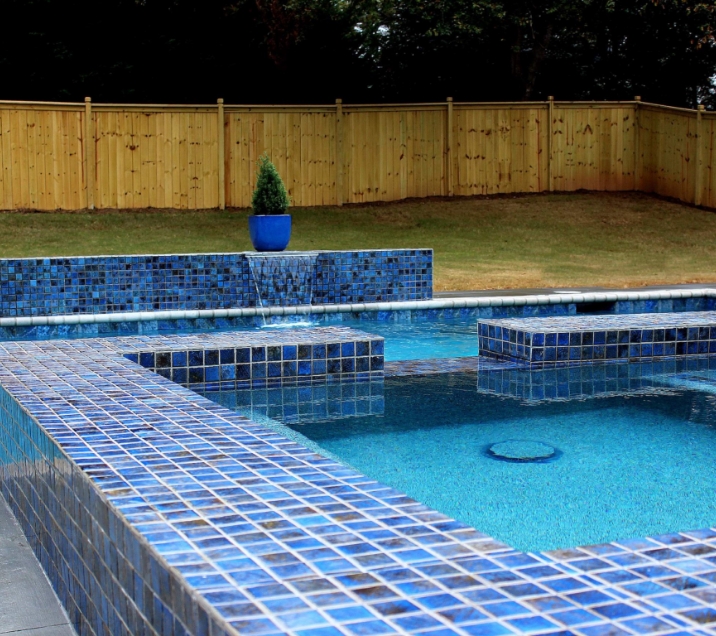 A swimming pool with a blue tile edge and two square step-like features in the water. The pool is surrounded by a wooden fence, and a potted plant sits on the edge. Green grass and trees are visible in the background.