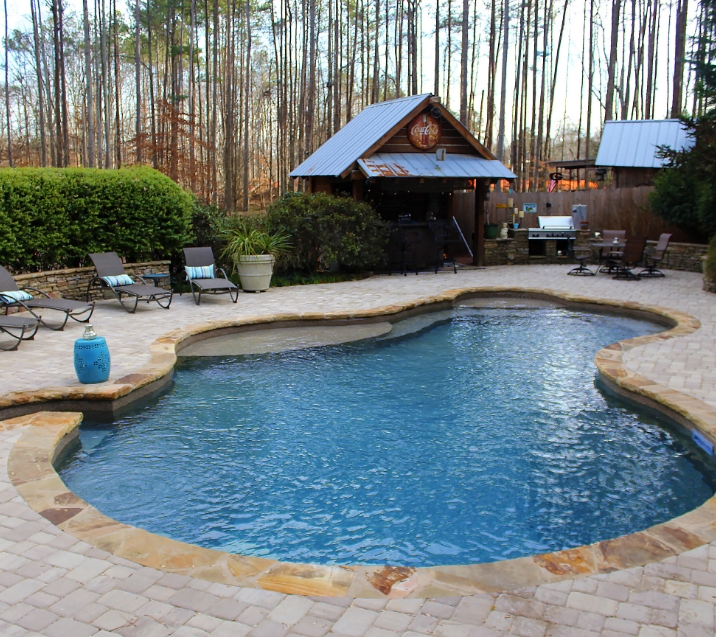 A backyard oasis featuring a freeform swimming pool with stone edging, surrounded by paver patio. Lounge chairs line the poolside, and a wooden cabana with a metal roof houses a bar and grill. Tall trees stand in the background.