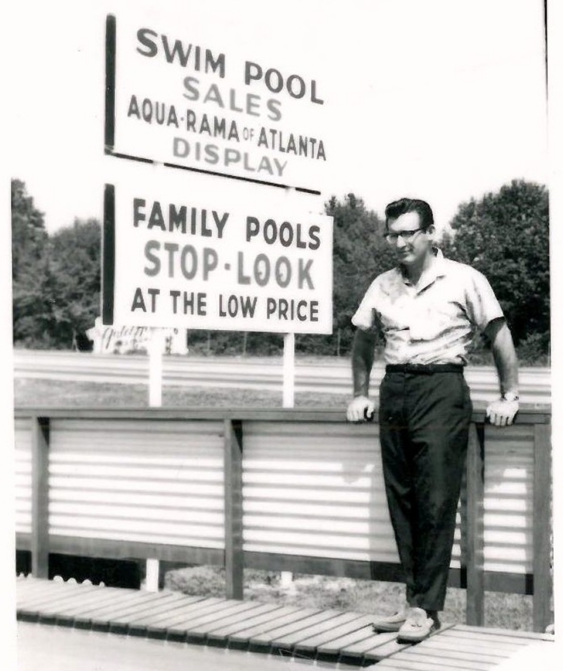 A man stands in front of signs advertising swim pool sales and family pool offers. The signs read Aqua-Rama of Atlanta Display and Stop - Look at the Low Price, with a wooded area in the background.