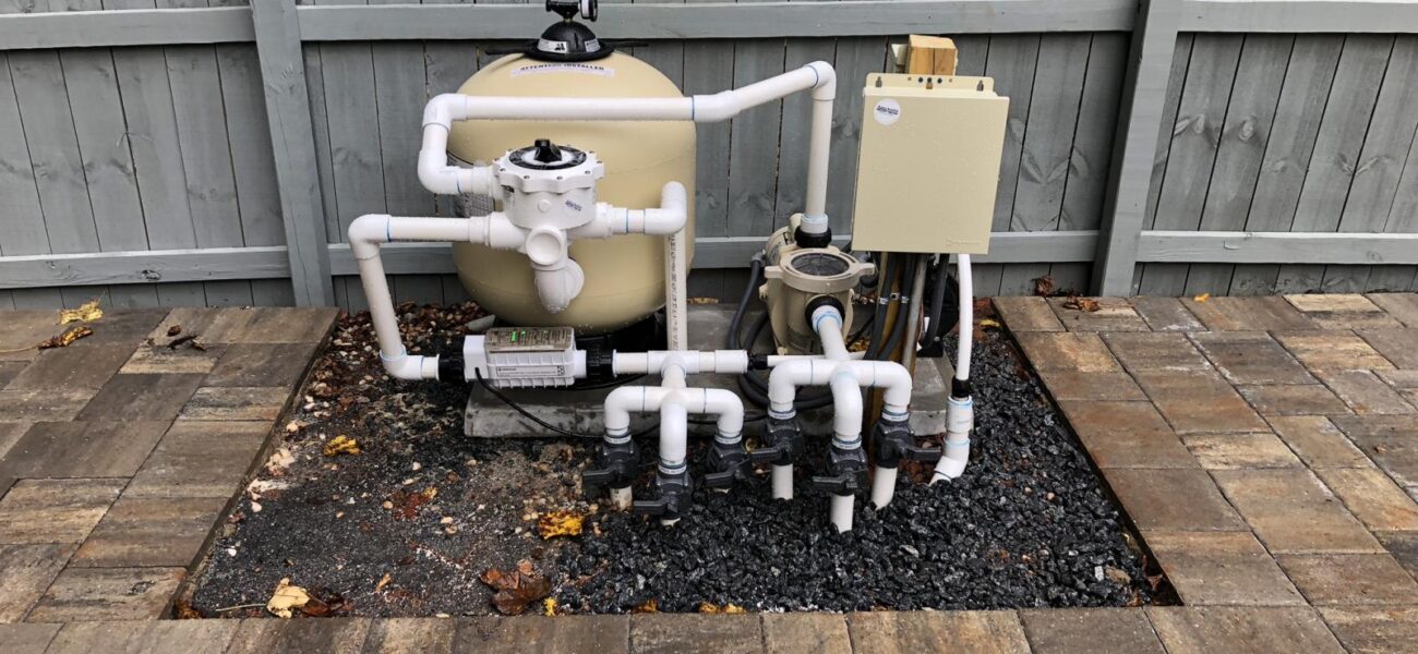 Pool equipment setup with a beige sand filter, white PVC pipes, and a control box, surrounded by a gravel bed. The area is bordered by interlocking stone pavers, set against a wooden fence. Dry leaves are scattered around.