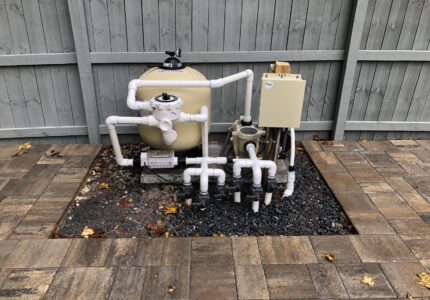 Pool equipment setup with a beige sand filter, white PVC pipes, and a control box, surrounded by a gravel bed. The area is bordered by interlocking stone pavers, set against a wooden fence. Dry leaves are scattered around.