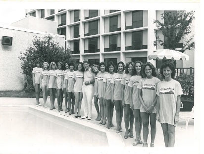 A black-and-white photo of a group of women standing in a line by a pool, all wearing matching shirts with text and shorts. They are outside a multi-story building with large windows. Trees and a patio umbrella are in the background.