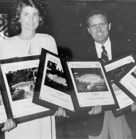 Two people stand side by side, each holding framed awards featuring photographs of landscaping projects. The woman wears a light blouse, and the man is in a suit and tie. The image is in black and white.