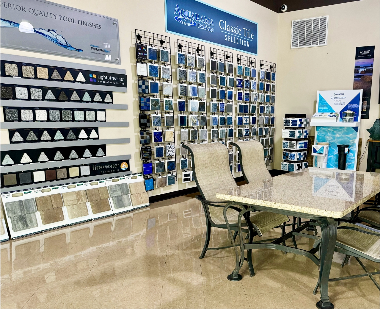 A showroom displaying various pool tiles and finishes. Sample boards of tiles are mounted on walls, with a table and chairs in the center. Signs label sections like Classic Tile Selection and Superior Quality Pool Finishes.