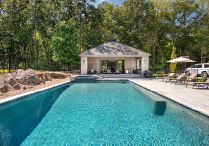 A rectangular outdoor pool with clear blue water is surrounded by a stone deck. Lounge chairs with umbrellas are on the right. A small pool house with seating is visible at the end. Trees and rocks border the area, creating a serene backdrop.