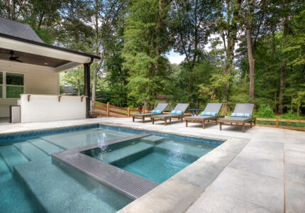 A serene outdoor swimming pool surrounded by lush trees. Four lounge chairs with striped pillows are lined up along the poolside. The house has a covered patio with a ceiling fan, offering a relaxing natural setting.