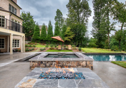 Outdoor patio with stone fire pit in foreground, overlooking a pool and lounge chairs. Brick house with chimney to the left and tall trees in the background under a cloudy sky.