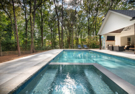 A modern backyard features a rectangular swimming pool with a stone deck, surrounded by tall trees. A covered patio with seating and two lounge chairs is visible on one side, while sunlight filters through the branches above.