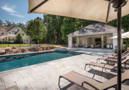 A backyard pool area with stone tiles features several lounge chairs under umbrellas. In the background, theres a white pool house with outdoor furniture. Trees and a large house with a black fence are visible in the distance.