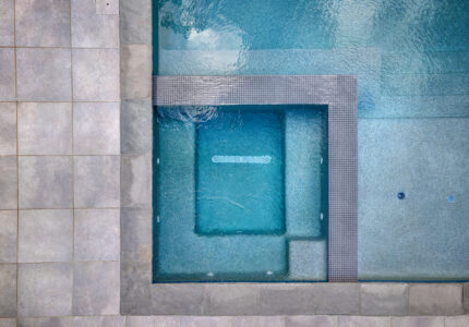 Aerial view of a modern swimming pool and hot tub. The hot tub, outlined with tiles, is connected to the pool. The surrounding pool deck is covered with large, square gray tiles, creating a sleek and contemporary look.