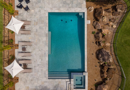 Aerial view of a rectangular swimming pool surrounded by a stone patio. Several lounge chairs and two umbrellas are placed on one side. A small spa is attached to the pool. Landscaping with rocks and green grass borders the area.