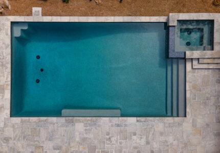 Aerial view of a rectangular swimming pool with a small attached square hot tub. The pool is surrounded by light-colored stone tiles, and theres a patch of brown earth visible at the top edge of the image.