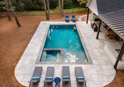 A rectangular swimming pool with an attached spa is surrounded by large tiles. There are six lounge chairs with striped pillows and a small round table beside the pool. A covered patio with seating is visible on the right. Trees and a fence are in the background.