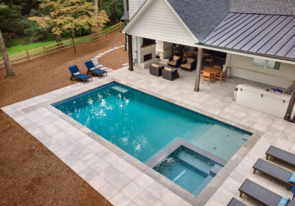 Aerial view of a modern backyard featuring a rectangular swimming pool with an attached hot tub. Surrounding the pool are several lounge chairs and a covered patio area with outdoor seating and a dining table, next to a house with a dark roof.