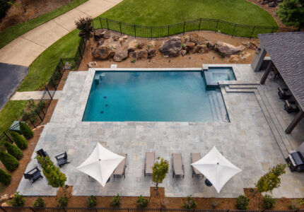 Aerial view of a rectangular swimming pool surrounded by a stone patio. The patio features three lounge chairs with white umbrellas. Adjacent to the pool is a hot tub. Landscaping with shrubs and a rock border is visible around the area.