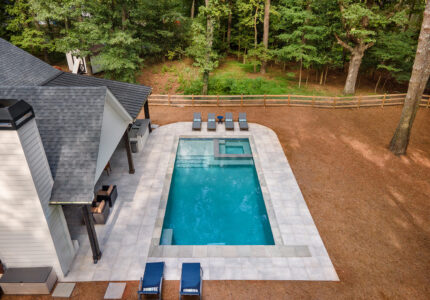 Aerial view of a rectangular backyard pool surrounded by a patio with several lounge chairs. The adjacent house has a covered outdoor sitting area. The background features a wooden fence and lush green trees.