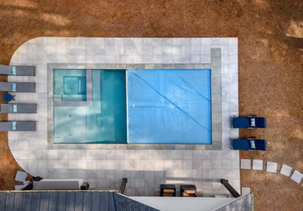 Aerial view of a rectangular swimming pool with a smaller hot tub section. The pool is partially covered. Surrounding the pool are lounge chairs with striped cushions. A curved path leads to the pool area.