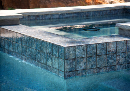 A close-up view of a square hot tub attached to a swimming pool, featuring blue and gray tiles. The water in the hot tub is clear, with gentle ripples, and the surrounding stonework is visible. Sunlight casts natural reflections on the waters surface.