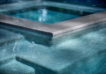 A serene image of a swimming pool with clear, blue water. The pool features a square, elevated spa section with tiled edges. Gentle ripples disturb the otherwise smooth surface, reflecting sunlight in patterns.