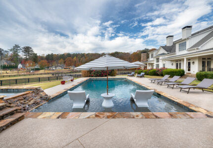 A luxurious backyard pool area with two white loungers and a round table shaded by a large umbrella. The pool features a waterfall edge. Several reclined chairs are on the deck, and a large house with chimneys is in the background under a partly cloudy sky.