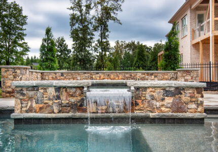 A tranquil pool with a stone waterfall feature flows into the foreground. Trees and a house are visible in the background, under a cloudy sky. The scene exudes a peaceful and serene atmosphere.