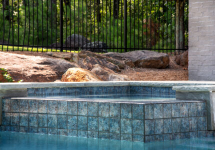 A swimming pool with a raised spa area located beside a brick wall. The pool is surrounded by natural rocks and backed by a black metal fence, with green trees visible in the background.