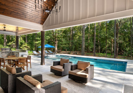 Outdoor patio with wicker furniture, a large rectangular pool, and a dining area under a wooden ceiling. The space overlooks a lush forest, with sunlight filtering through the trees. A chandelier hangs above, adding elegance to the open area.