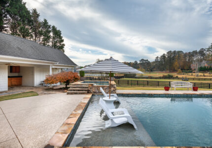 Outdoor pool area with two white lounge chairs partially submerged in the water. A striped umbrella provides shade. A stone patio surrounds the pool, and there’s a view of a fenced grassy field with trees in the background.