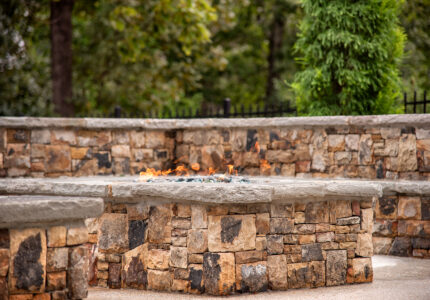 A stone fire pit with small flames surrounded by a circular stone bench in a garden setting. Behind are trees and a metal fence. The scene conveys a cozy outdoor atmosphere.